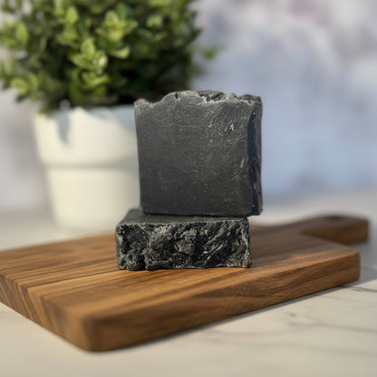 Two pieces of black soap on a light surface with greenery in the background